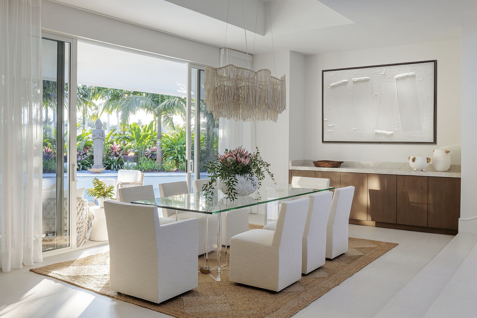 Glass dining table with sculptural chairs framed by sheer drapery and a tropical view